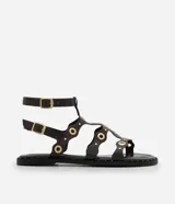 Sandalia plana tipo gladiadora color negro, confeccionada en poliuretano. Presenta tiras onduladas con recortes ahuecados y detalles de ojalillos y tachas metálicas doradas. Cuenta con doble tira ajustable en el tobillo con hebillas metálicas.