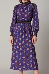 Vestido midi violeta con estampado floral, cuello alto con volados, mangas largas abullonadas y cintura elastizada con detalle de franja horizontal.