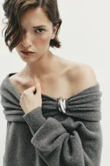 Jersey de punto color gris oscuro, con escote recto tipo off-shoulder, manga larga y detalle de aplique metálico en el hombro. Presenta acabados en rib en puños y ruedo.