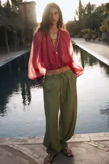 Pantalón ancho de tiro alto color beige claro, con cintura elástica, cordón ajustable y pinzas delanteras. Presenta bolsillos frontales y bolsillos de plastrón en la espalda. Incluye un cinturón del mismo tejido con apliques metálicos.