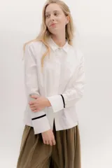 Camisa blanca de corte masculino con cuello clásico y botones dorados. Los puños tienen un detalle de cinta de terciopelo negro. La espalda presenta un juego de cintas en contratono y el ruedo trasero es redondeado con tablón.