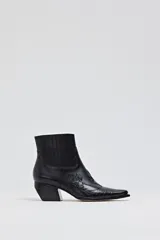 Bota corta de cuero negro con textura de cocodrilo en la puntera y detalles de costuras decorativas. Presenta un taco cuadrado inclinado y paneles laterales elásticos.