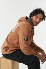 Campera con capucha de felpa color marrón, con estampado tipográfico en la espalda, bolsillo tipo cargo en la manga y puños elásticos.