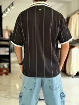 Camiseta deportiva blanca de manga corta con rayas verticales negras, cuello polo en V de color negro con ribete blanco, y puños negros. Cuenta con estampado frontal con la palabra "TOKIO" en grande, además de detalles gráficos y texto en el pecho.