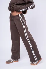 Pantalón de jogging color marrón, de corte recto y holgado, con cintura elástica y doble franja lateral blanca. Presenta una etiqueta pequeña con la marca en la parte inferior de la pierna.