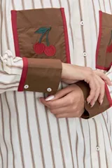 Camisa de corte oversize con estampado de rayas verticales, cuello clásico y cierre frontal de botones. Presenta dos bolsillos frontales en contraste con bordados de tréboles, además de puños y cuello en color sólido con detalles en amarillo.