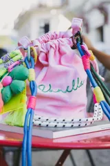 Cartera tipo bucket verde con base estructurada, cordón de ajuste rosa y azul, y llaveros colgantes de flor azul y corazón rosa.