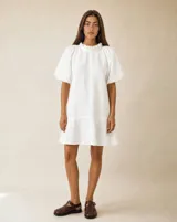 Vestido corto color blanco, confeccionado en lino 100% natural, con silueta amplia y relajada, cuello fruncido con volados y mangas cortas abullonadas.