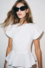 Top de popelina blanca con cuello redondo, mangas cortas abullonadas y detalle de abertura en la espalda con lazada. Presenta bajo en evasé tipo peplum y cierre lateral con cremallera oculta.