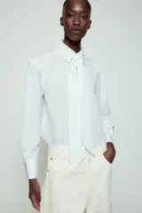 Camisa blanca de cuello solapa y manga larga, con detalle de lazada en el mismo tejido en la parte frontal y cierre con botones dorados.