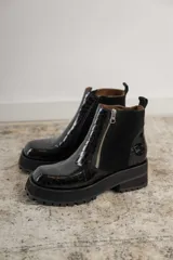 Botas cortas negras con plataforma dentada, confeccionadas en una combinación de cuero con textura de reptil y paneles de gamuza. Presentan un cierre frontal metálico y puntera cuadrada.