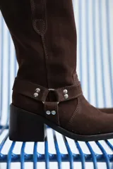 Bota de caña media confeccionada en cuero gamuzado color chocolate. Presenta un diseño de estilo western con taco cuadrado de 7 cm y una correa decorativa en el tobillo con herrajes metálicos plateados.