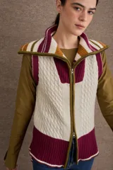 Chaleco de lana con cierre frontal completo y cuello alto. Presenta un diseño de bloques de color en tonos crema, bordó y detalles en mostaza, con una combinación de tejido trenzado y acanalado. El diseño es más largo en la parte posterior y cuenta con aberturas laterales.