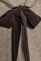 Chaqueta de punto con capucha, manga larga y cierre frontal con cremallera. Presenta un diseño en color beige con un lazo decorativo a contraste en color marrón oscuro en la zona del cuello.