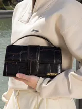 Bolso Le Grand Bambino de Jacquemus confeccionado en cuero de ternera con estampado efecto cocodrilo en color marrón oscuro. Presenta una silueta estructurada, solapa frontal con cierre magnético y tira vertical, asa superior rígida y correa ajustable y desmontable. Incluye detalles metálicos dorados con el logotipo de la marca en el frente.