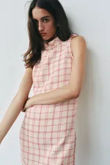 Vestido corto sin mangas de tejido texturizado con diseño de cuadros en tonos crudo y fucsia. Presenta cuello subido, trabillas decorativas con botones dorados en los hombros y cierre lateral con cremallera oculta.