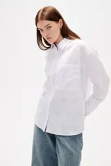 Camisa de algodón blanca con estampado de corazones formados por pequeños cristales de strass. Presenta cuello clásico, cierre frontal con botones, bolsillo en el pecho y mangas largas con puños abotonados.
