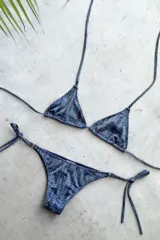 Bikini de dos piezas con estampado que simula tela vaquera o denim con brillo. El corpiño es de estilo triángulo con tiras finas ajustables y apliques de argollitas. La bombacha es vedetina, también de atar en los laterales con apliques de argollitas.