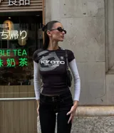 Top de manga larga con diseño superpuesto, que combina una remera de manga corta negra con estampado gráfico frontal y mangas largas blancas debajo. Presenta un ajuste al cuerpo y estilo deportivo.