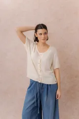 Cardigan tejido de manga corta color beige, con escote redondo amplio y abotonadura frontal con botones al tono. Presenta un calce relajado y terminaciones en rib en la cartera y el ruedo.
