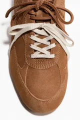 Calzado deportivo de gamuza suave en color marrón, con diseño de perfil bajo, collarín acolchado y doble juego de cordones en tonos marrón y blanco.