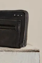 Billetera de cuero color negro con acabado gastado, diseño alargado, cierre perimetral de cremallera y tirador de cuero trenzado. Cuenta con bolsillo exterior con cierre y marca grabada en el frente.