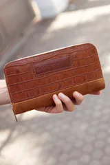 Billetera mediana marrón de cuero con textura croco y detalles en gamuza. Tiene cierre perimetral y logo Austera grabado en el frente.