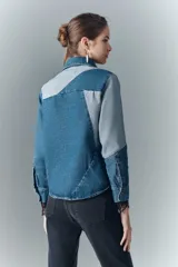 Campera recta de denim rígido con combinación de recortes en tonos de azul. Cuenta con bolsillos de solapa en el pecho y botones metálicos. Las mangas tienen puños con encaje negro.