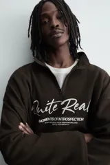 Sudadera de corte boxy fit en tejido de algodón con textura acanalada, color marrón oscuro. Presenta cuello subido con cierre frontal de cremallera y estampado frontal con texto en contraste.