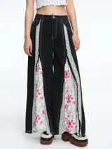 Pantalón de corte wide leg en color negro, con paneles laterales insertados que combinan encaje y tela estampada con motivos florales. Presenta costuras a la vista en contraste y diseño de silueta amplia.