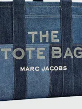 Cartera tote bag Marc Jacobs modelo The Traveler Tote, confeccionada en denim azul con el nombre de la marca estampado en el frente. Cuenta con doble asa superior.