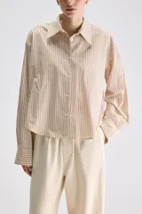 Camisa de calce relajado confeccionada en algodón, con diseño de rayas verticales en tonos beige y blanco. Presenta cuello clásico, cierre frontal con botones, puños abotonados y largo ligeramente cropped.