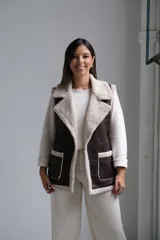 Chaleco de cuero sintético color chocolate con interior y bordes de corderito beige. Presenta solapas amplias, dos bolsillos frontales con detalle de corderito y corte recto.