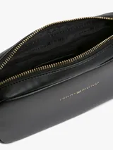 Cartera tipo bandolera Tommy Hilfiger de color negro, con acabado liso, diseño compacto y esquinas redondeadas. Cuenta con cierre de cremallera superior, inscripción y logotipo de la marca en bajo relieve dorado en el frente, y una correa cruzada ancha ajustable de jacquard a rayas en blanco y negro con la marca escrita.