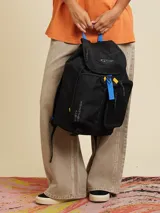 Mochila negra de poliéster con detalles en azul y amarillo. Tiene un bolsillo frontal con cierre, un bolsillo lateral de red y el logo de Rusty Worldwide estampado.