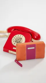 Billetera mediana de cuero sintético con diseño bicolor en naranja y fucsia. Cuenta con cierre perimetral, correa de muñeca extraíble y placa metálica decorativa con logo.