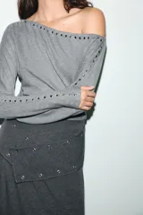 Camiseta de cuello barco y manga larga tipo murciélago, confeccionada en tejido de punto gris melange. Presenta detalle de tachas metálicas a lo largo de los hombros y las mangas.