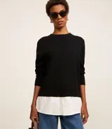 Suéter negro de punto con cuello redondo y mangas largas. Tiene un dobladillo superpuesto de camisa blanca.