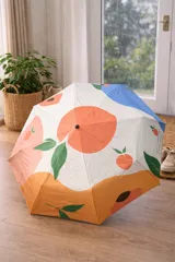 Paraguas plegable con estampado frutal de duraznos sobre fondo blanco y detalles en azul en la parte inferior. Incluye correa de mano negra.