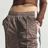 Pantalón Adidas Adibreak de corte holgado, confeccionado en poliéster reciclado con estampado animal print tipo jirafa en tonos marrón y blanco. Cuenta con bandas laterales blancas con líneas negras y botones a presión, además del logo clásico de la marca.
