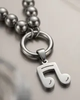 Set de joyería en acero quirúrgico compuesto por un collar con dije de nota musical y aros tipo bolita. El collar presenta una cadena con eslabones finos y una sección de esferas metálicas que conectan con un aro central del cual cuelga el dije. Los aros son de diseño minimalista en forma de esfera con cierre de presión.