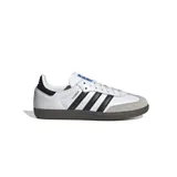 Championes Adidas modelo Samba OG, de diseño clásico con exterior de cuero blanco, tres tiras laterales negras, puntera de gamuza gris y suela de goma color marrón.