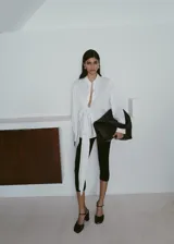 Camisa blanca de manga larga con cuello camisero, cierre frontal de botones y un llamativo detalle de nudo drapeado en el lateral delantero.