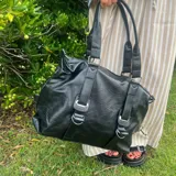 Bolso negro de cuero sintético con doble asa de hombro y correas decorativas frontales con hebillas metálicas. Incluye cierre superior y bolsillo interior.