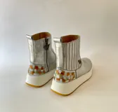 Botas cortas de cuero plateado, con diseño híbrido que combina la estética texana con una suela chunky blanca y detalle de arte lateral creado por Carolina De Cunto.