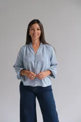 Blusa de tela tipo denim liviano en tono azul claro, con cuello mao, cierre frontal de botones y mangas largas con puños. Presenta un delicado bordado floral en todo el frente y las mangas.
