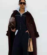 Trench coat largo de gamuza sintética con cuello peaked, cierre frontal cruzado con botones y cinturón ajustable en la cintura. Presenta mangas largas con reguladores en los puños y forro interior.