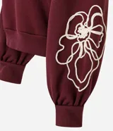 Buzo de algodón color bordó con cuello redondo acanalado, mangas largas con puños ajustados y terminación inferior elástica. Presenta un diseño de bordado floral en cuerda de color crudo en el frente y en una de las mangas.
