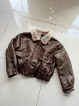 Chaqueta tipo aviador de cuero sintético en color marrón, con interior y cuello de corderito sintético en tono crema. Presenta cierre frontal, bolsillos con solapa y botones a presión, y puños y cintura con elástico acanalado.