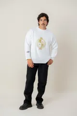 Buzo de tejido de punto color blanco, de corte holgado y cuello redondo. Presenta un bordado central en hilo dorado con el escudo nacional y un pequeño detalle bordado en la manga derecha con los colores de la bandera uruguaya.